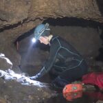 A person wearing a black suit and hard hat with a flashlight on it bends over a circular wall of a small cave.