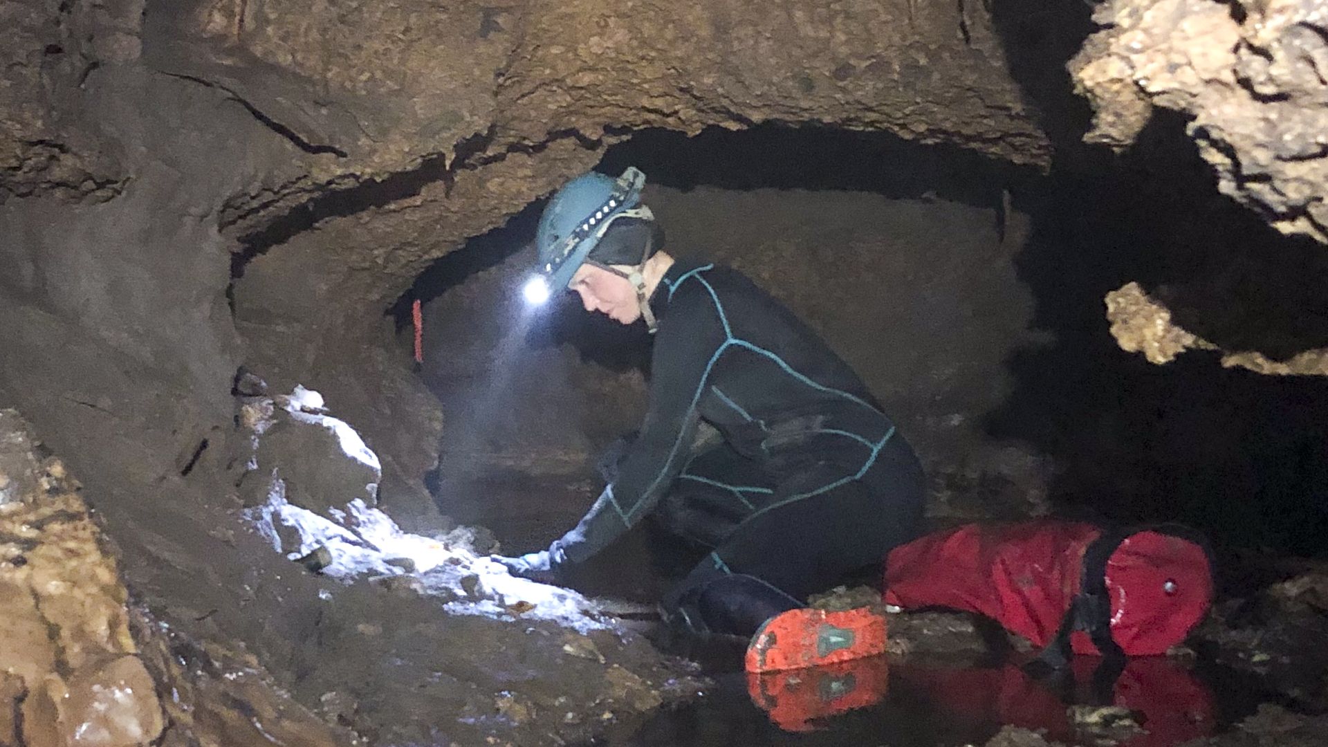 A person wearing a black suit and hard hat with a flashlight on it bends over a circular wall of a small cave.