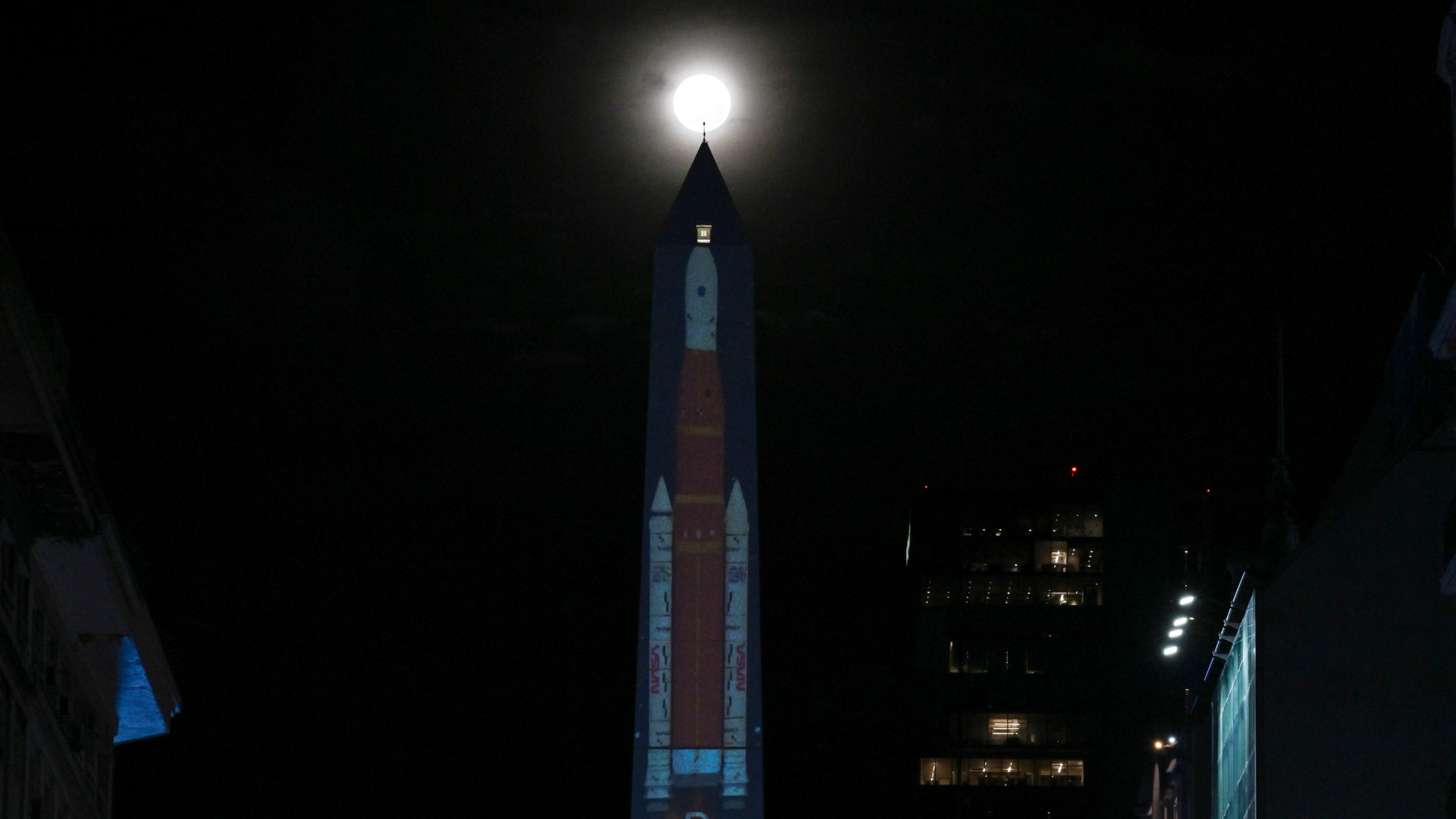 A large orange and white rocket is projected from the side of the obelisk at night surrounded by buildings. The moon appears to be shining on the top of the monument.
