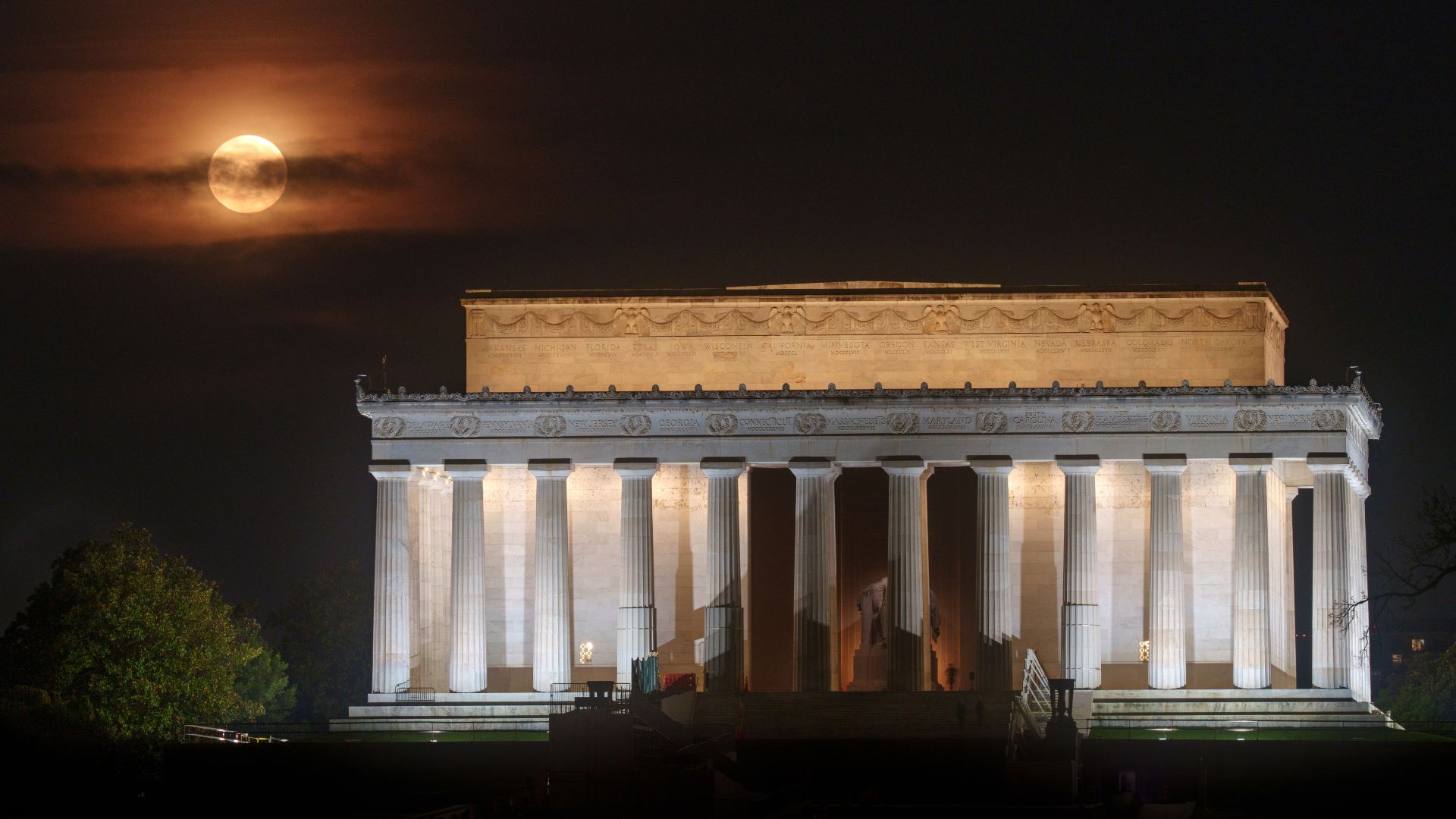 A full yellow moon is depicted in a cloudy night sky, shining over the Lincoln Memorial in Washington DC.
