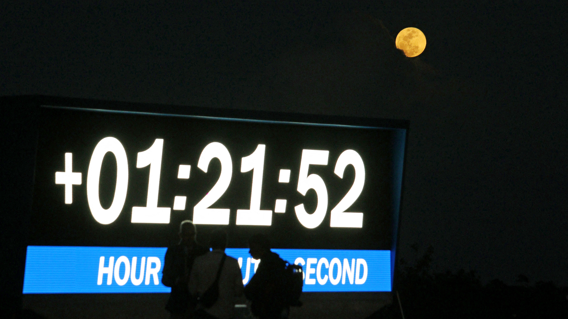 A full moon shines in the night sky above the launch pad at the Kennedy Space Center, with people standing in front.
