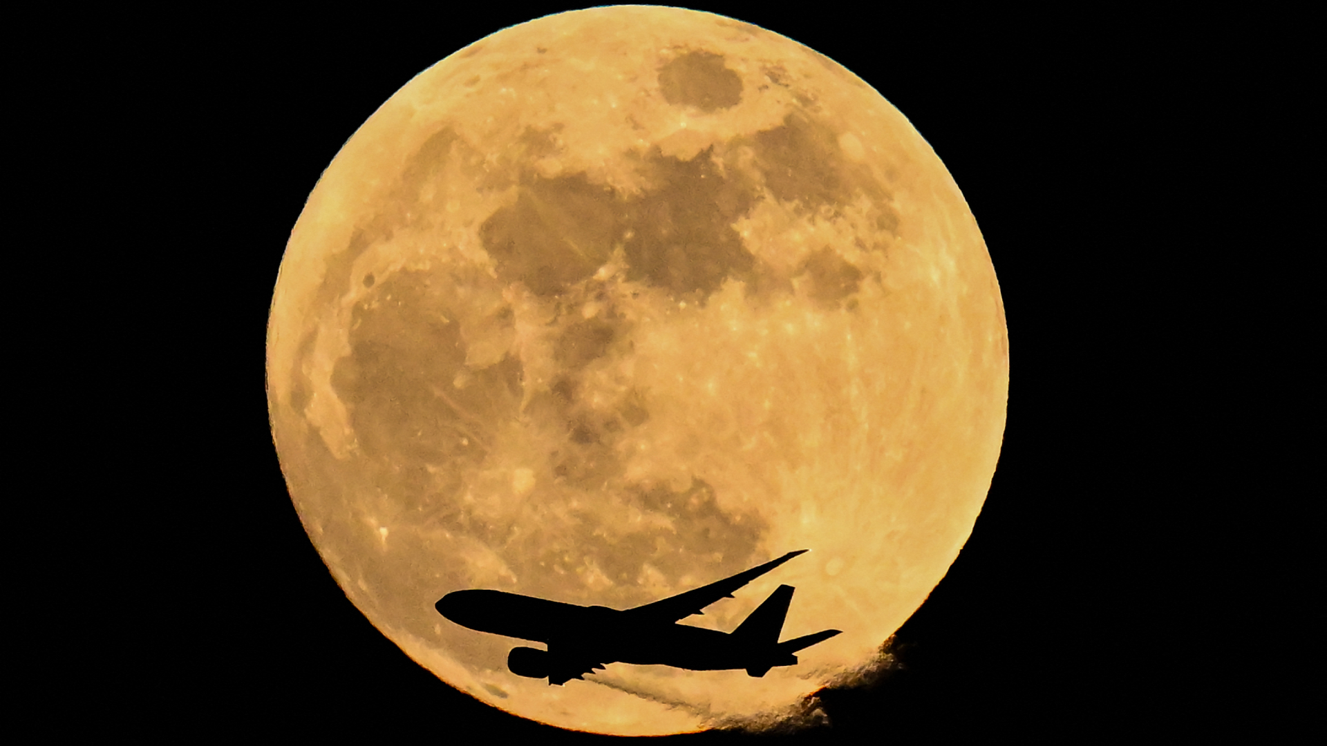 A full yellow moon shines in the black sky as the image of a passenger jet passes below it.