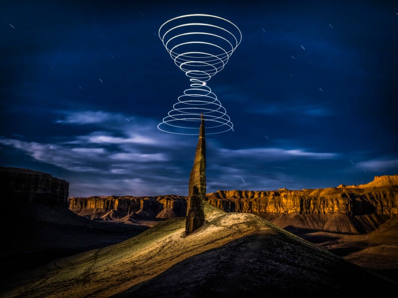 A tall rock peak stands in the desert landscape at night, with light trails spiraling above and below its summit; distant cliffs lit up under a starry sky.