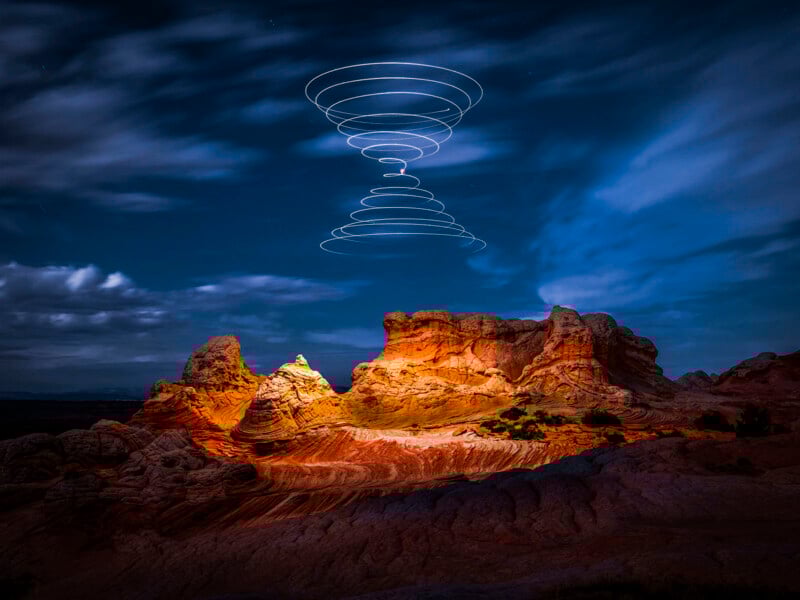 The red rock formations are lit up under the night sky, and there are winding white trails above, forming a mysterious spiral pattern. Some clouds are drifting across the sky, and the unique landscape is strikingly unique.
