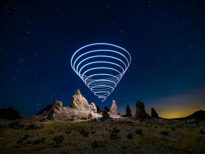 The rocks are shaped under the starry night sky, and there are spiral-shaped light trails, creating a bright light in the sky. The landscape looks dry because of the bare vegetation.