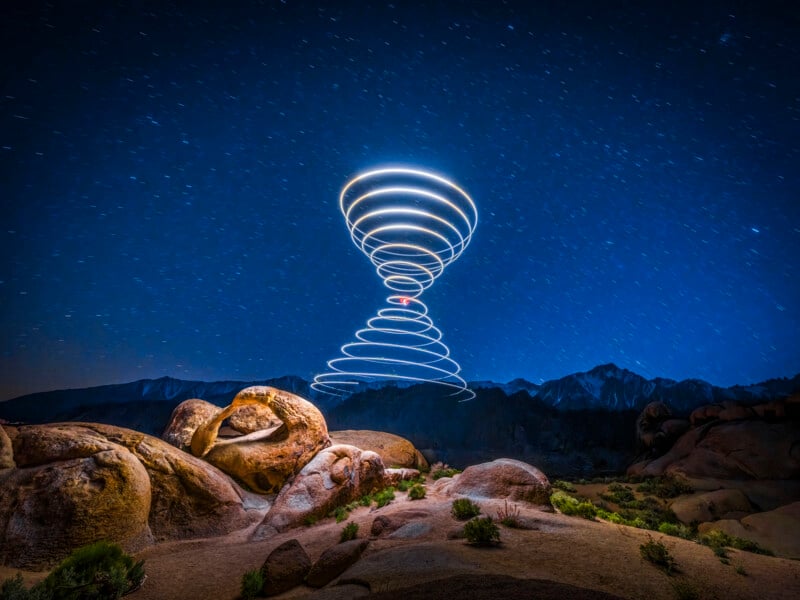 The night sky over a rocky landscape shows swirling light trails forming a storm-like pattern, with scattered stars in a deep blue sky and mountains in the background.