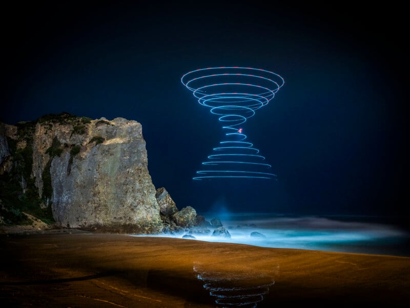 A rocky beach at night with a swirling image of blue and red light drawn in the sky, possibly from a long exposure aerial photograph, shown on the wet sand below.