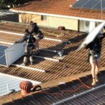 Men in dark clothes stand on a terracotta-tiled roof pulling out solar panels.