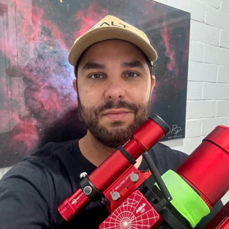 A bearded man wearing a cap and dark T-shirt is shown holding a red telescope and smiling at the camera in front of a nebula painting hanging on the wall.
