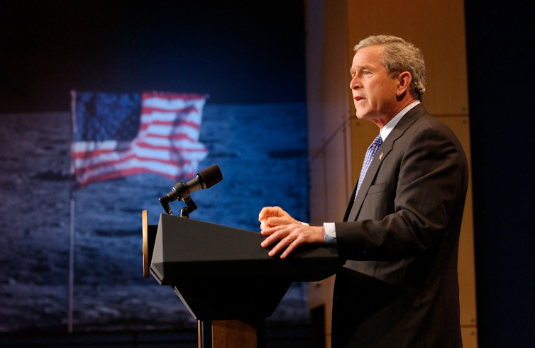 George W. Bush standing on a podium with an image of the American flag on the moon in the background.