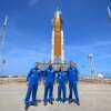 The four crew members of Artemis II, dressed in blue jumpsuits, stand in front of the rocket.